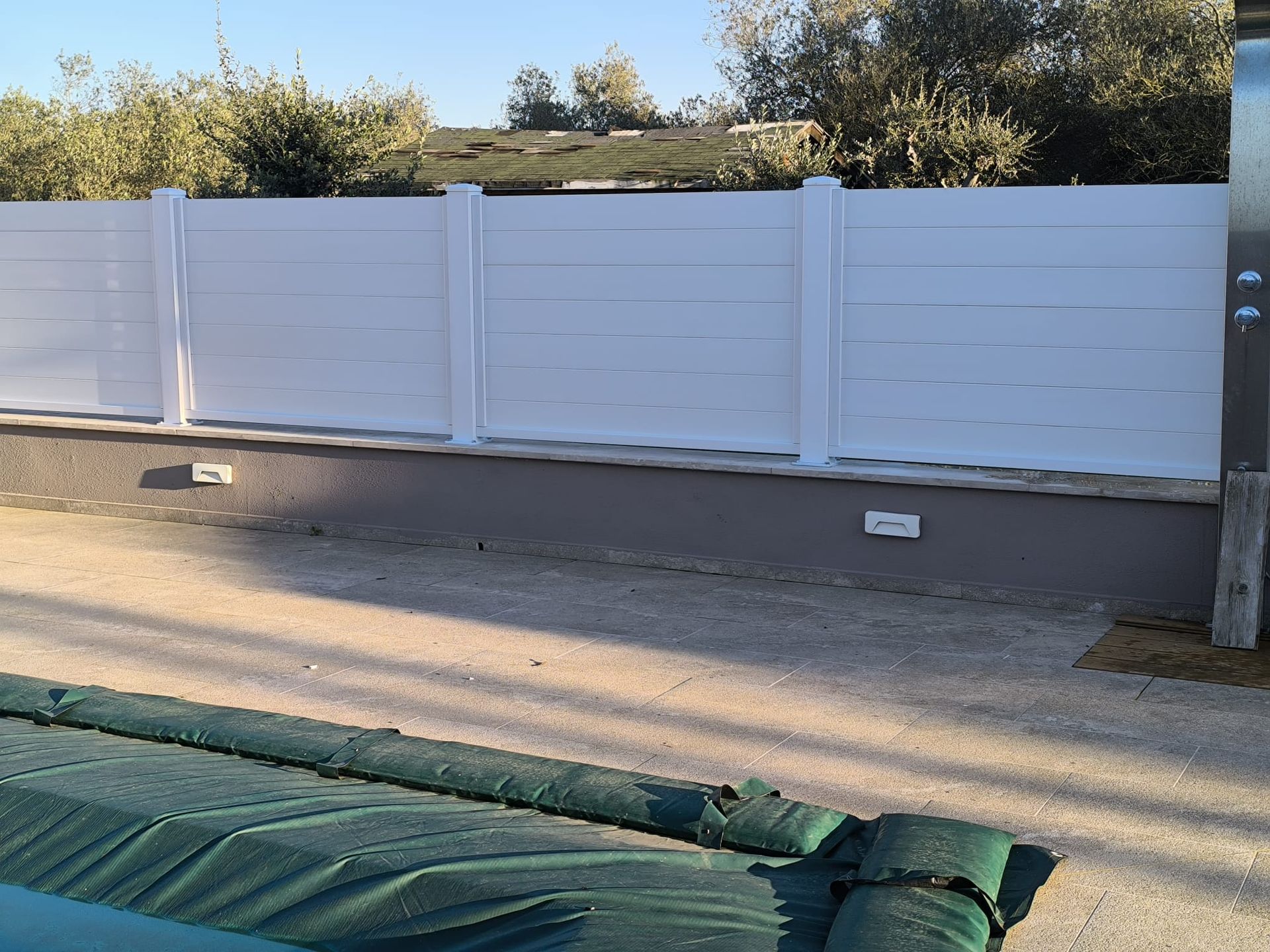 Recinzione bianca a doghe orizzontali con pali, terreno in ghiaia, copertura verde per la piscina. Alberi sullo sfondo.