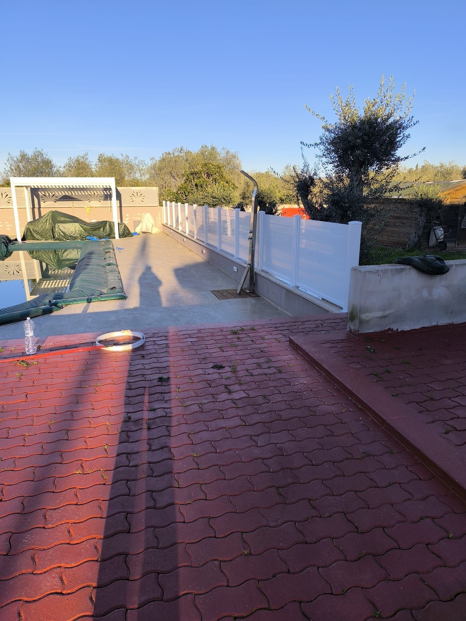 Un patio in mattoni rossi conduce a una recinzione bianca, a una piscina e ad alberi sotto un cielo azzurro e terso.
