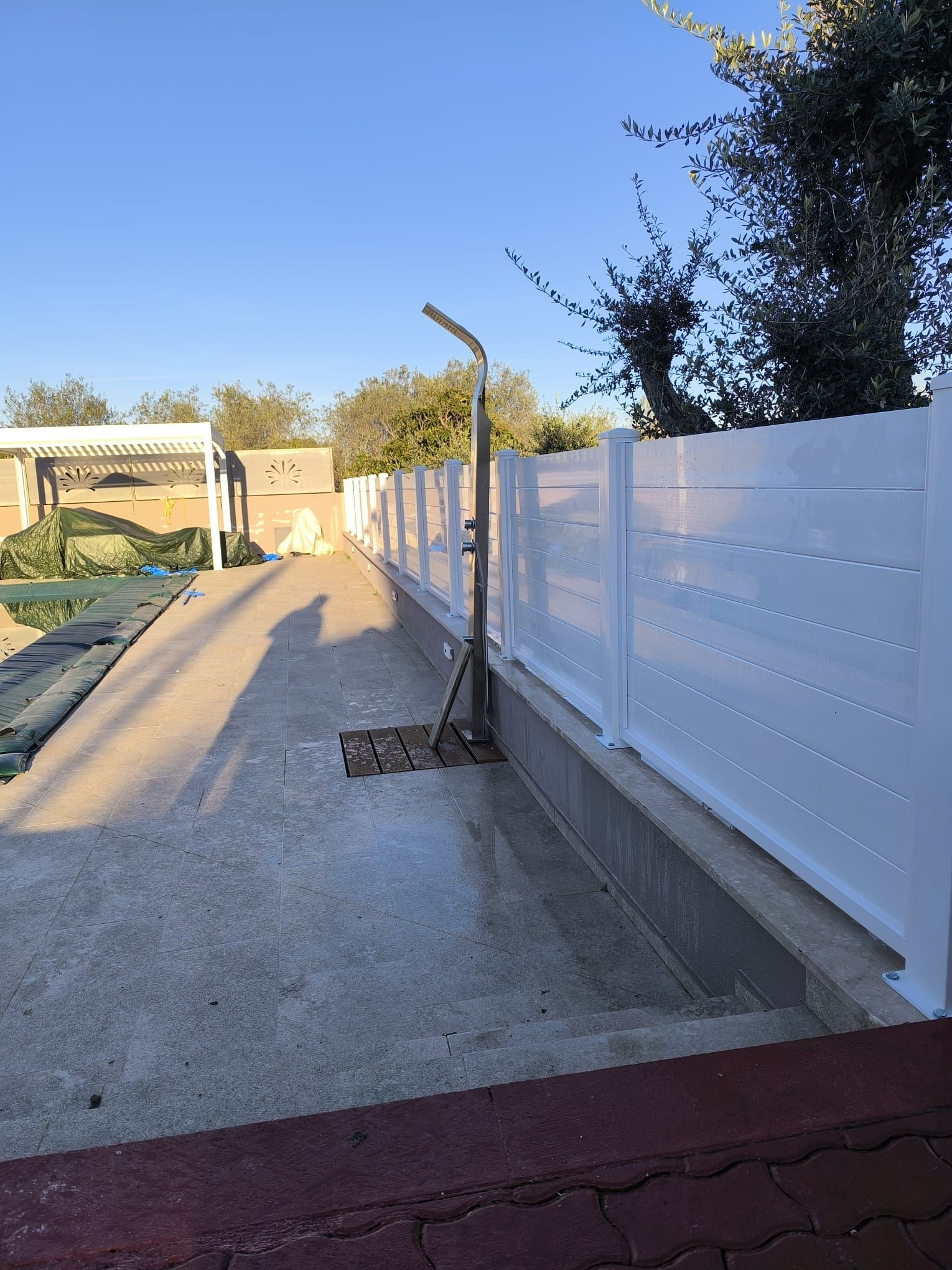 Doccia esterna accanto a una staccionata bianca e a un patio in cemento. Luce solare intensa.