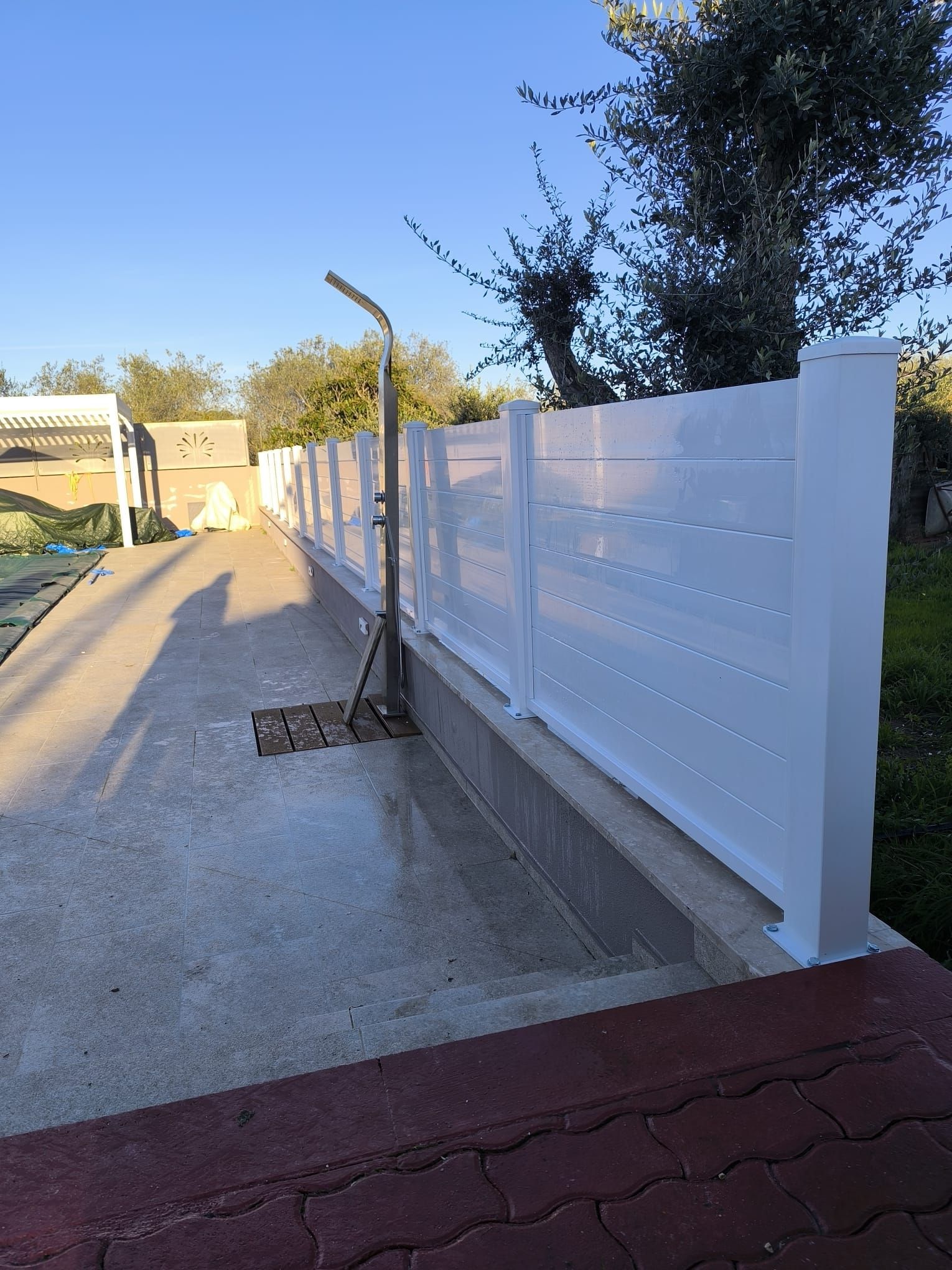 Recinzione bianca con listelli orizzontali lungo un vialetto in cemento, con doccia esterna e gradino rivestito di piastrelle rosse.