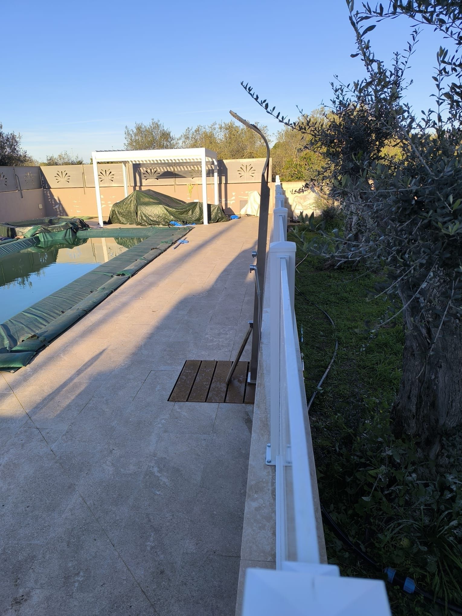 Terrazza in cemento con piscina, recinzione, doccia e una pergola sullo sfondo. Giornata di sole.