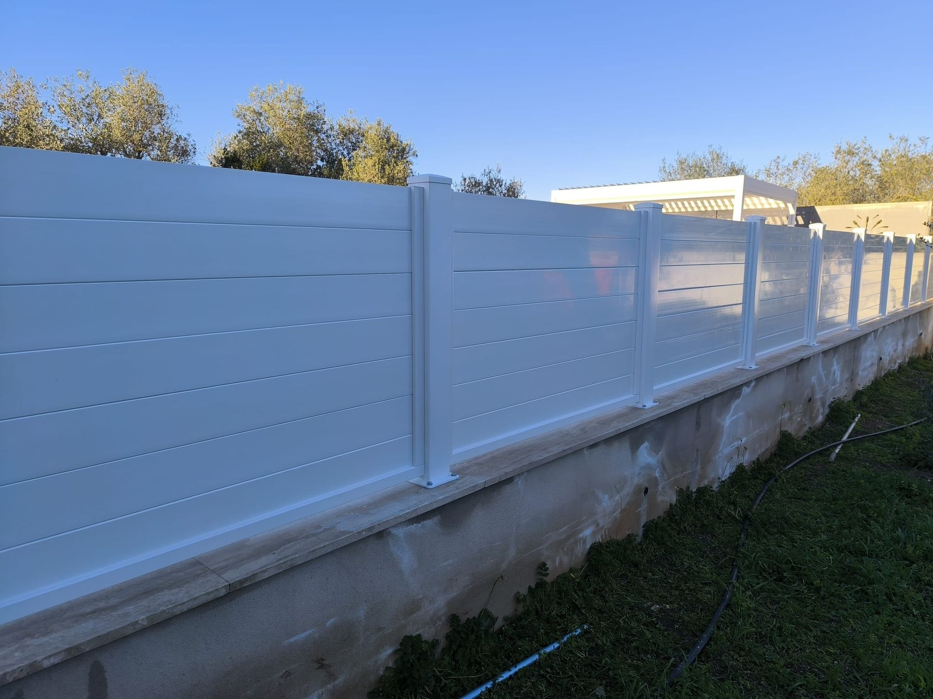 Recinzione bianca con listelli orizzontali su base di cemento, che si estende lungo un prato sotto un cielo azzurro.