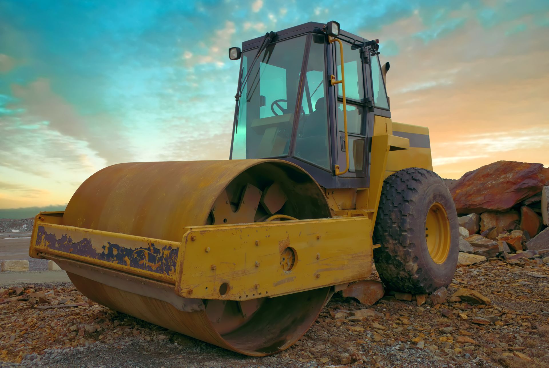 A large yellow road roller parked on rocky ground at sunset. A large yellow road roller parked on rocky ground at sunset.