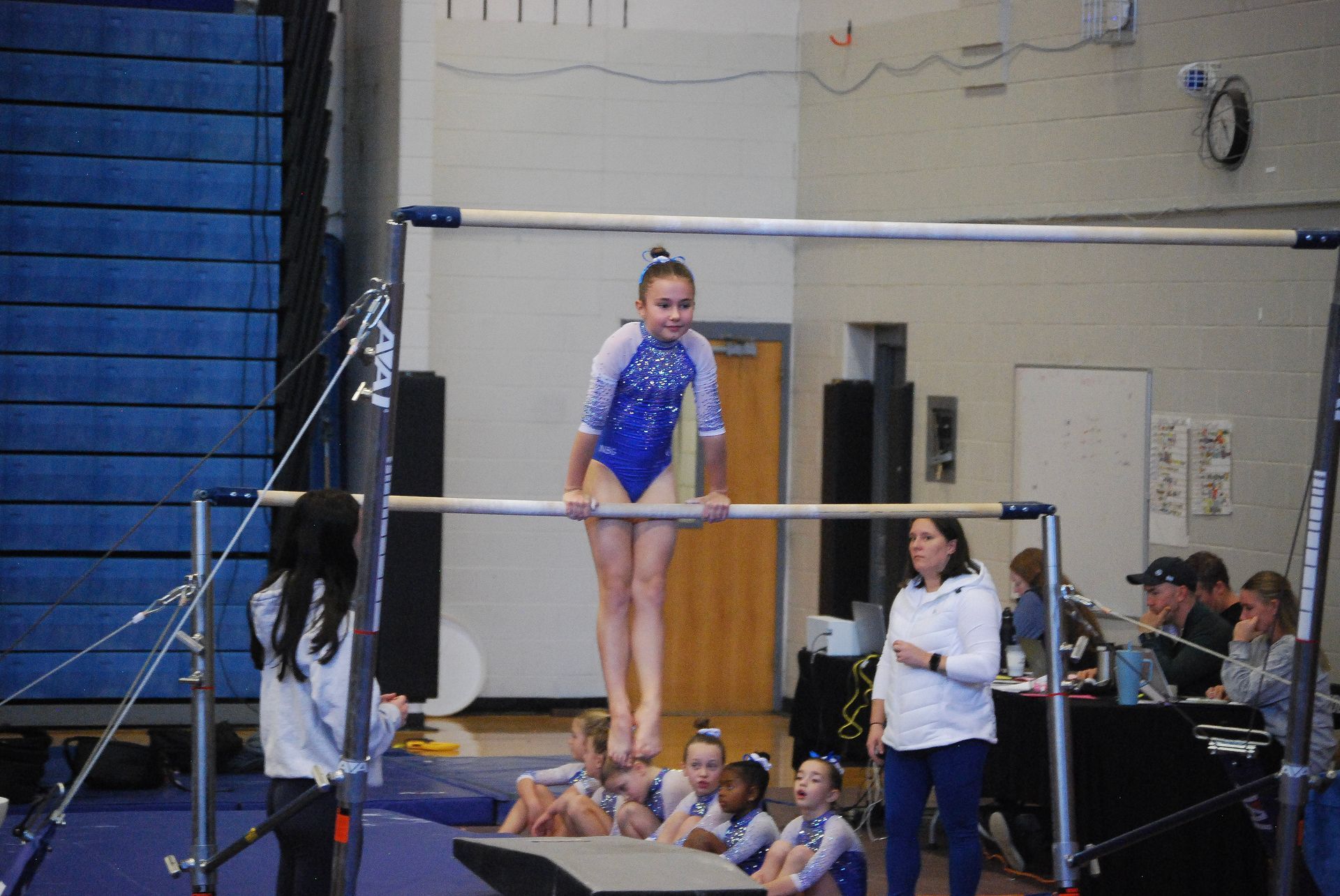 Girl in blue leotard on uneven bars, legs extended. Gymnastics competition indoors.