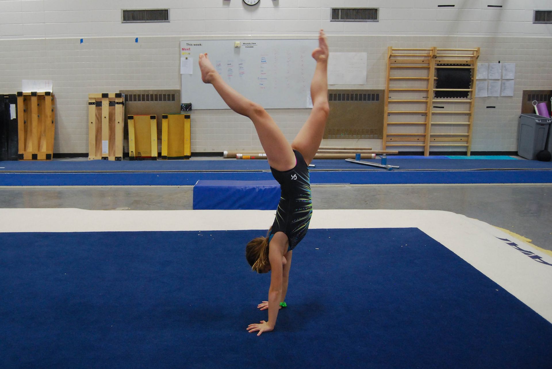 Gymnast doing a handstand on a blue mat, legs split in a V-shape. Gymnastics gym setting.