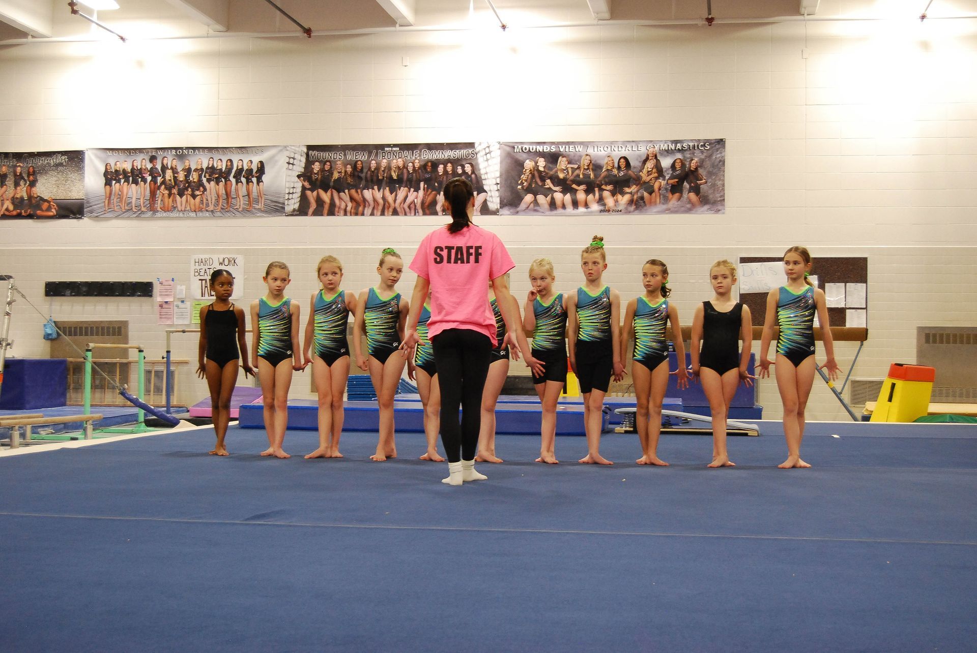 Gymnasts in leotards lined up on a blue mat, facing an instructor wearing a pink