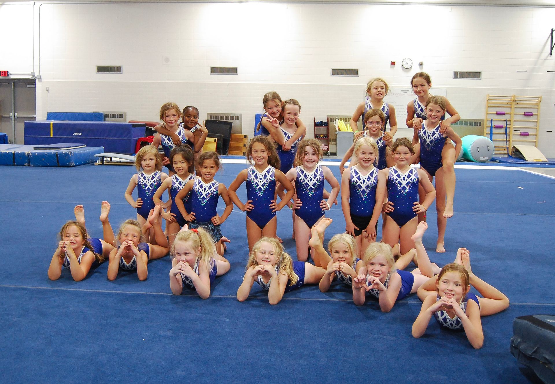 Gymnastics team posing on a blue mat, wearing blue leotards, smiling in a gymnasium.