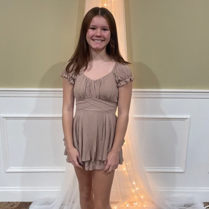 Teen girl in tan romper smiles, standing in front of a decorated wall.