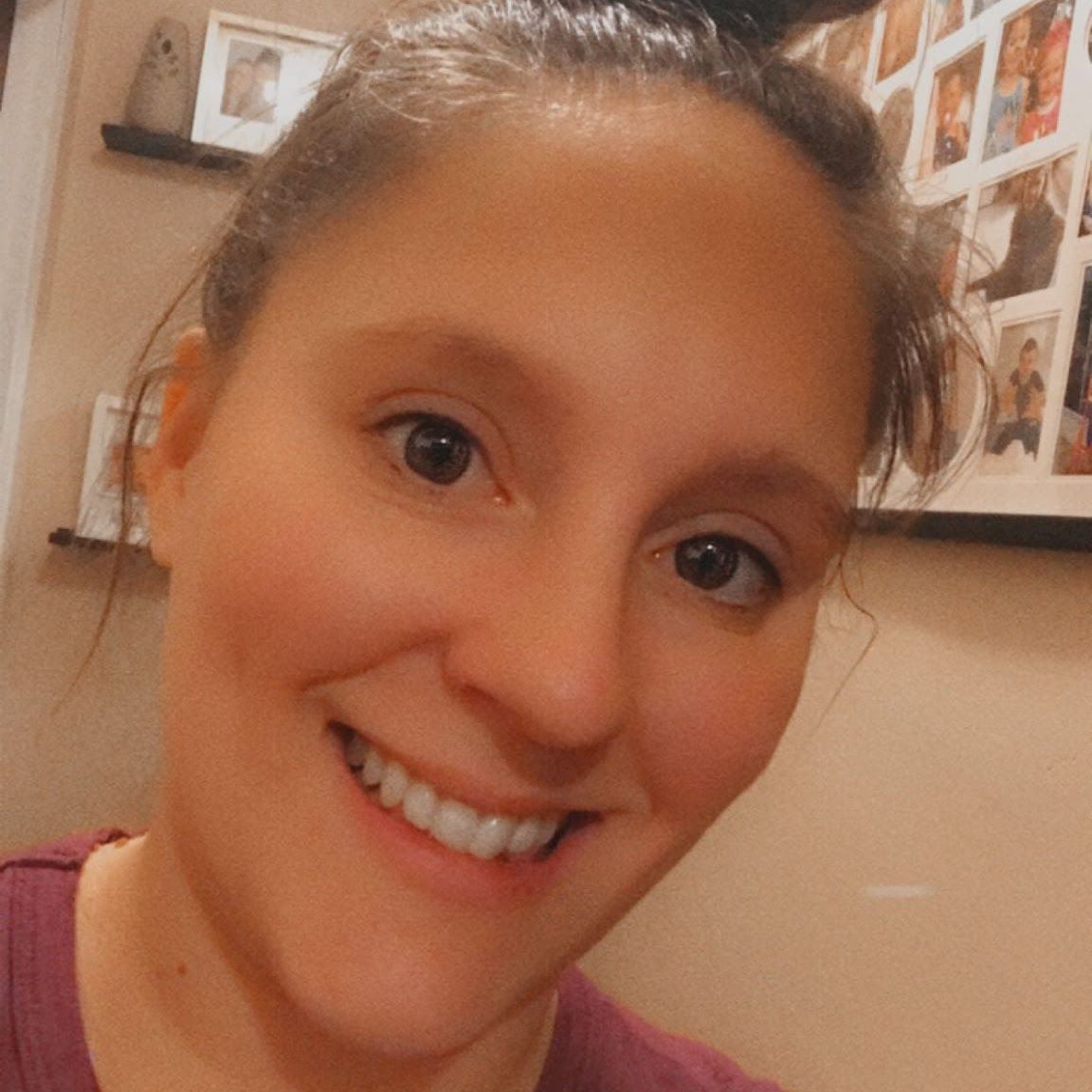 Woman with brown hair in a bun, smiling. Wearing a maroon shirt indoors, shelves and photos in background.