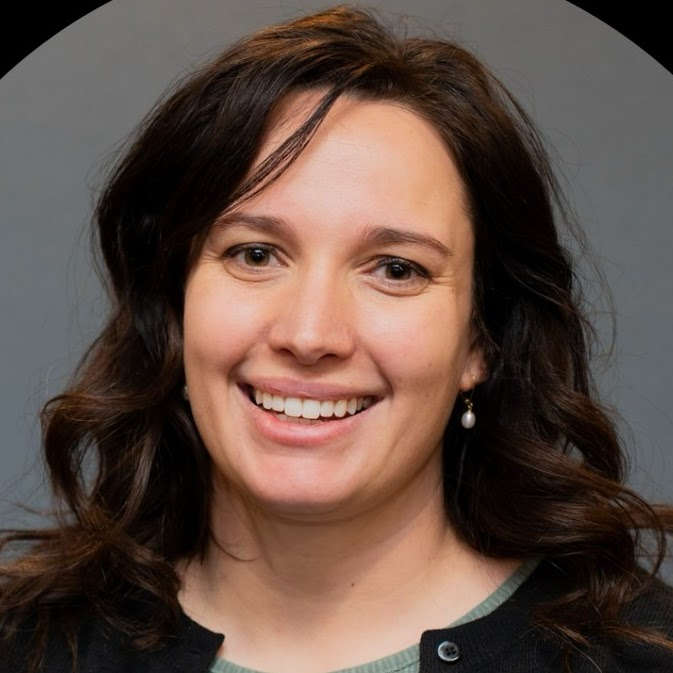 Woman with dark hair smiles, wearing a black cardigan and green shirt, in front of a gray background.