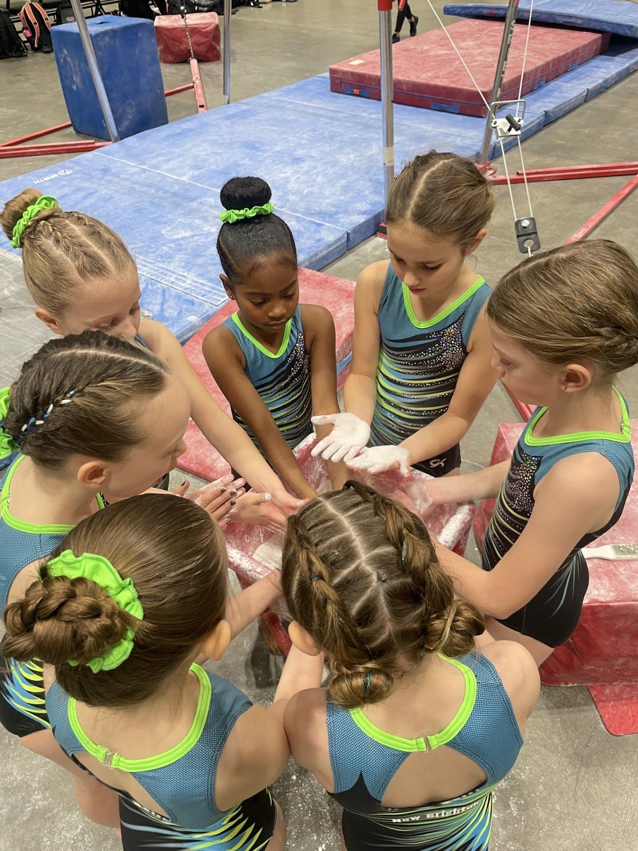 Young gymnasts in matching leotards put hands together, covered in chalk, at a gym.