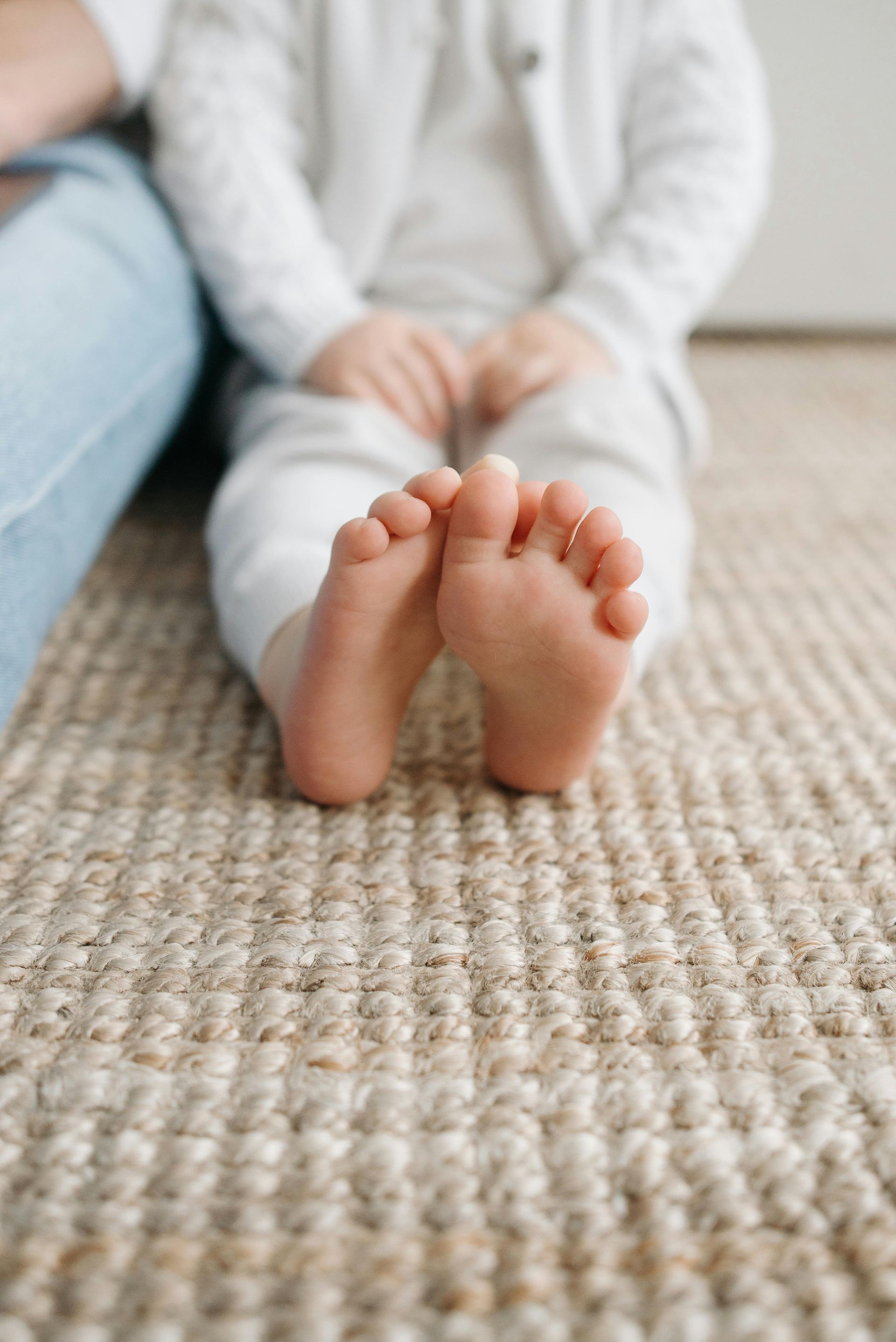 Bare feet of a child sitting on a tan textured rug, legs and arms out of focus.