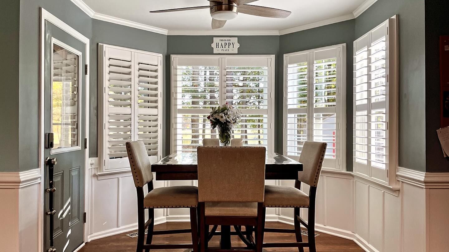A dining nook with a dark wood table, upholstered chairs, white window shutters, and sage green walls.