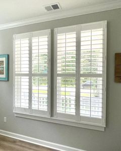 Two white plantation-style shutters installed over a large, side-by-side window set against a light gray wall.