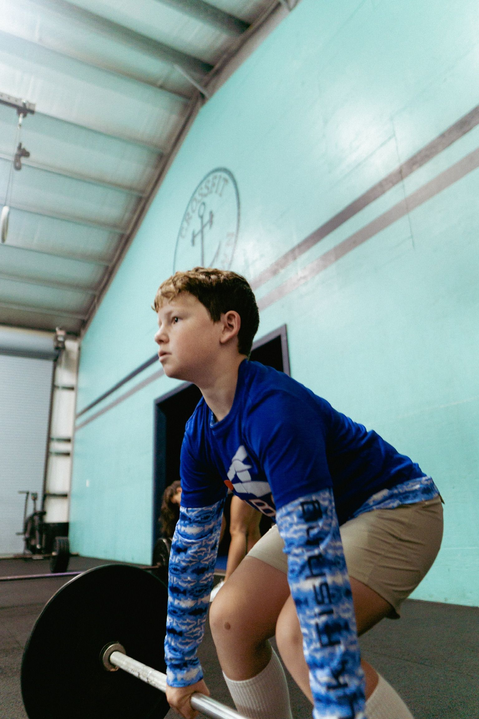 A person wearing a blue long-sleeve shirt and tan shorts bends over to lift a barbell at Crossfit Anchor Down