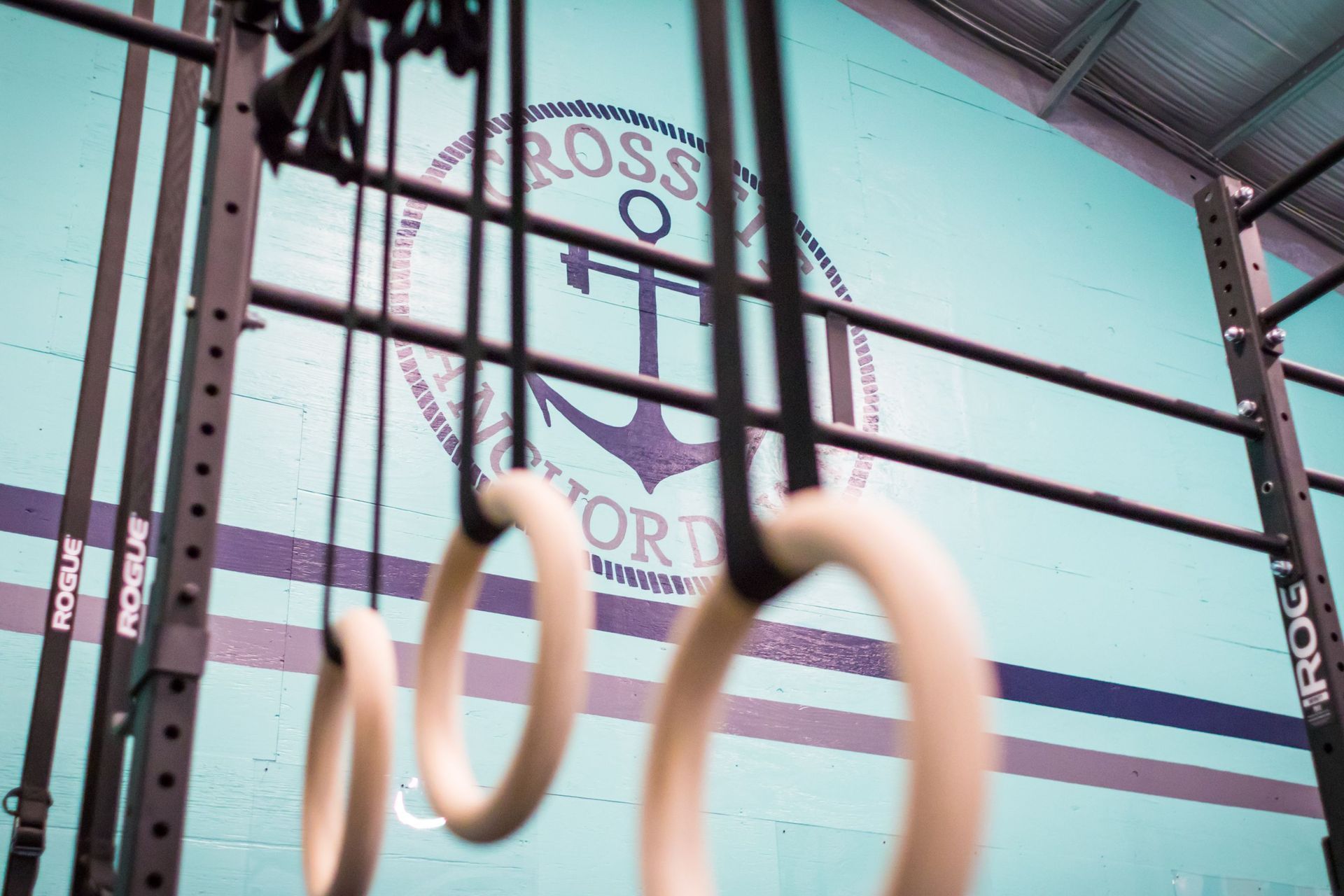 Wooden gymnastics rings hang in the foreground of a gym with a blue wall featuring the CrossFit Anchor Down logo.