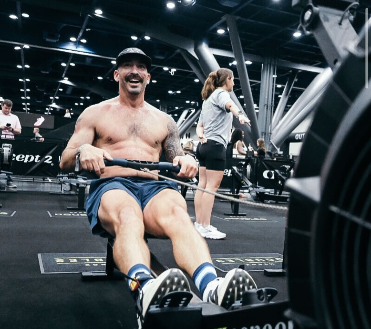 A smiling person shirtless in a baseball cap performs a rowing exercise on a machine inside a gym.