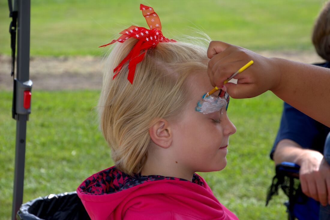 A little girl with a red bow in her hair is getting her face painted.