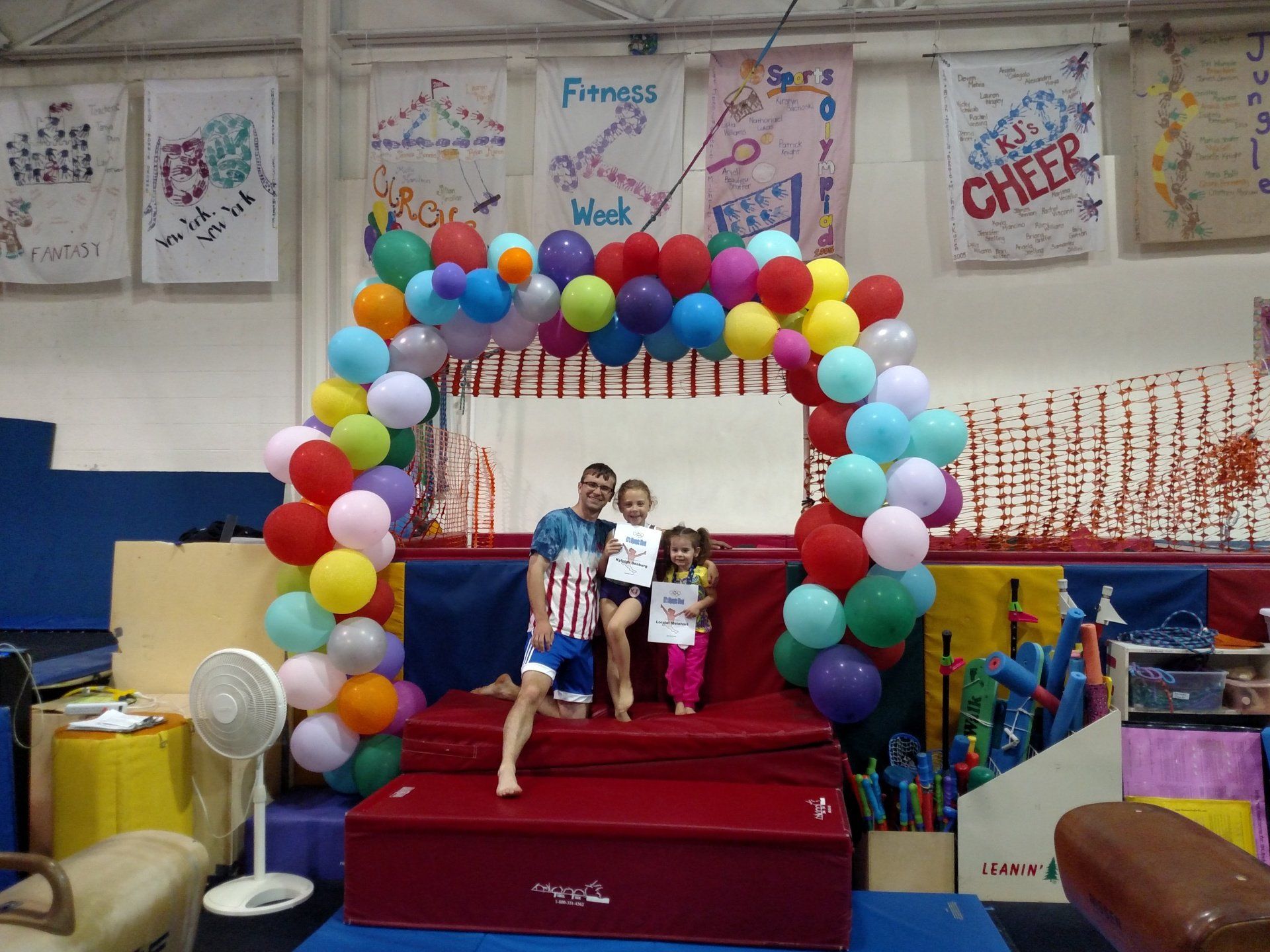 A man and two children are standing under a colorful balloon arch in a gym.