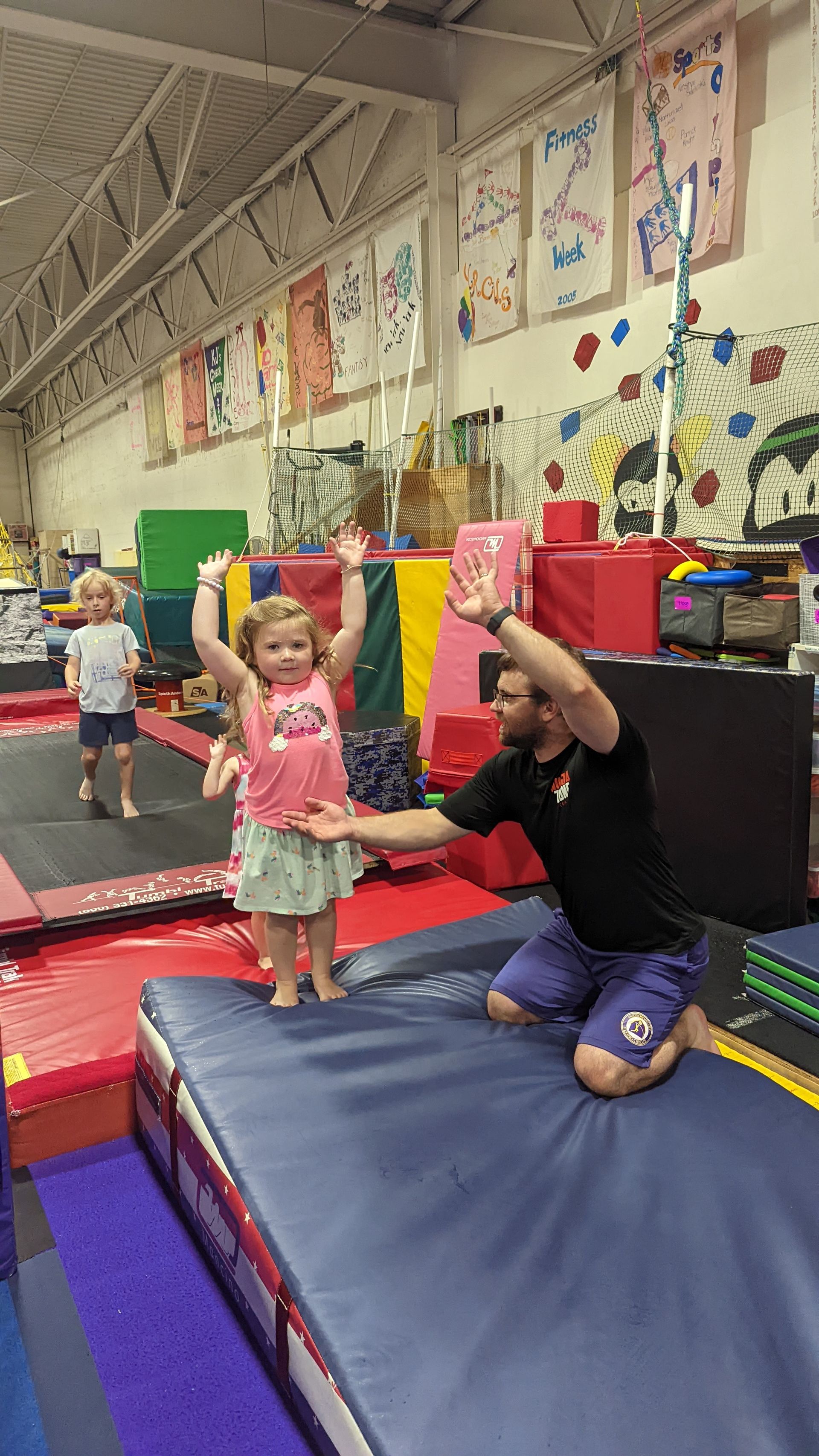 A man is kneeling down next to a little girl on a mat in a gym.