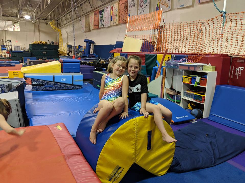 A boy and a girl are sitting on a foam roller in a gym.