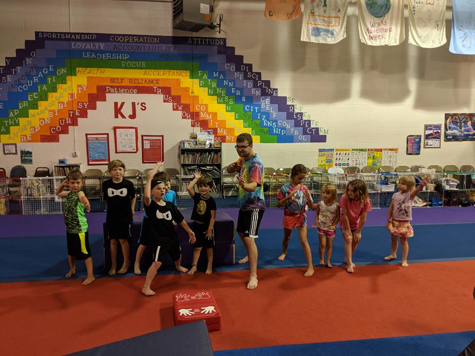 A group of children are standing in front of a rainbow wall in a gym.