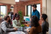 Group of colleagues in office, man presenting data on tablet, brick wall backdrop.