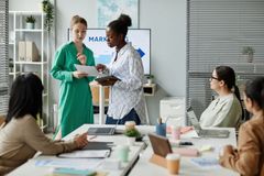 Two women present paperwork to a diverse office meeting. Whiteboard and laptops on table.