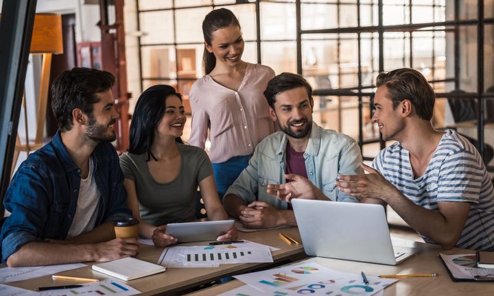 A diverse group of coworkers collaborating around a table with laptops and documents, smiling in a modern office.