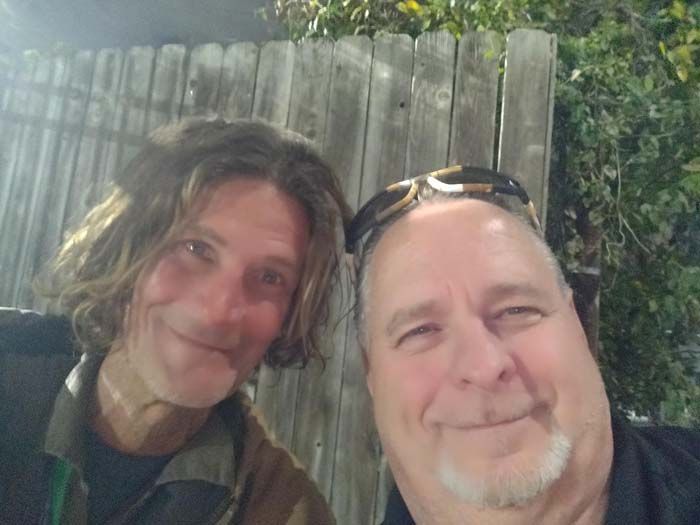 Two men are posing for a picture in front of a wooden fence.