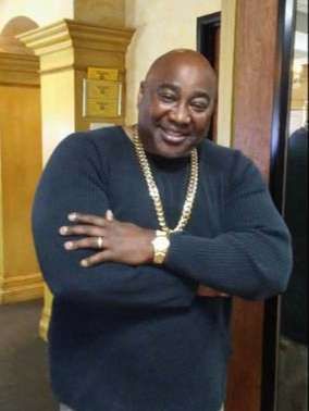 A man wearing a black sweater and a gold chain is standing with his arms crossed.