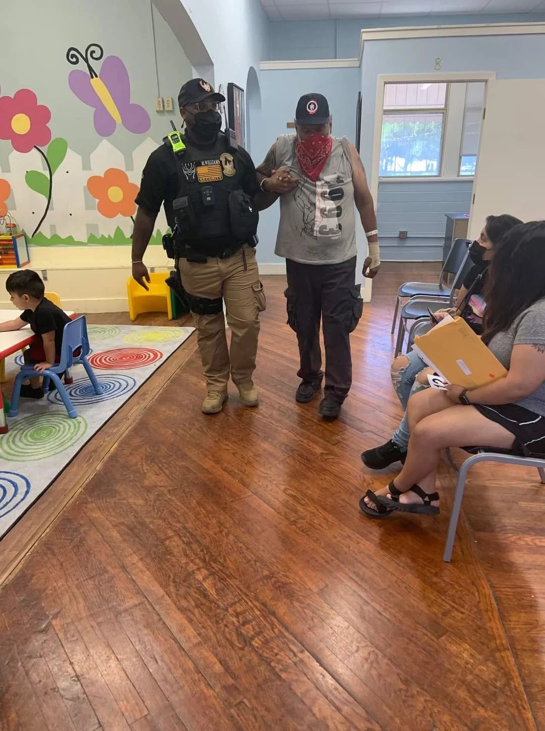 A man in a bandana is walking with a police officer in a room with people sitting in chairs.