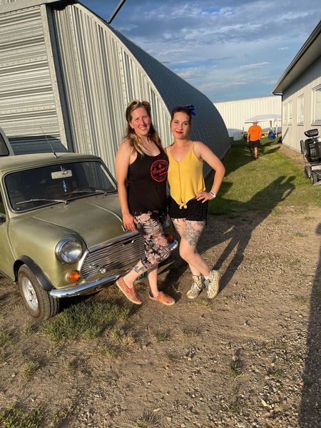Two women are standing next to each other in front of a car.