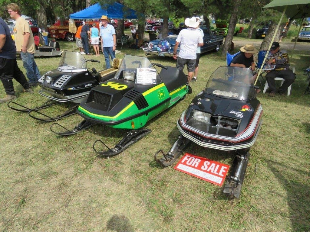 Three snowmobiles are parked in a grassy field with a for sale sign on the back.