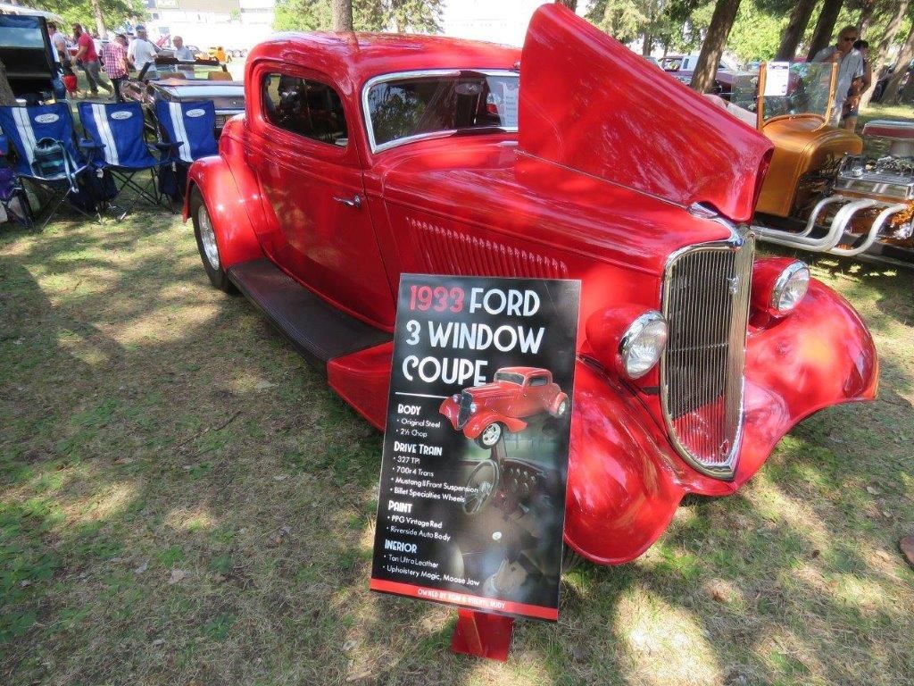 A red ford 3 window coupe is parked next to a sign.