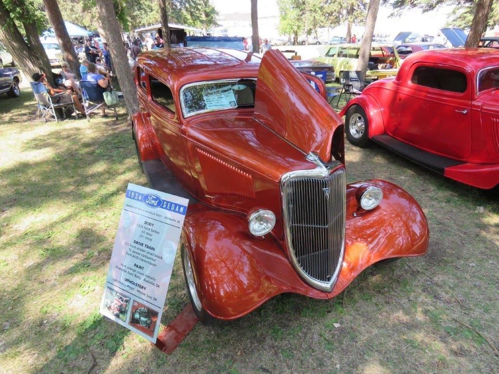 A red car with the hood up is parked in the grass at a car show.