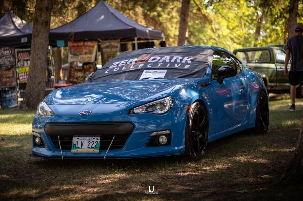 A blue car is parked in a park with trees in the background.