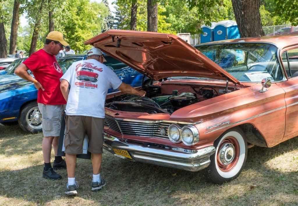 Two men are looking under the hood of an old car at a car show.
