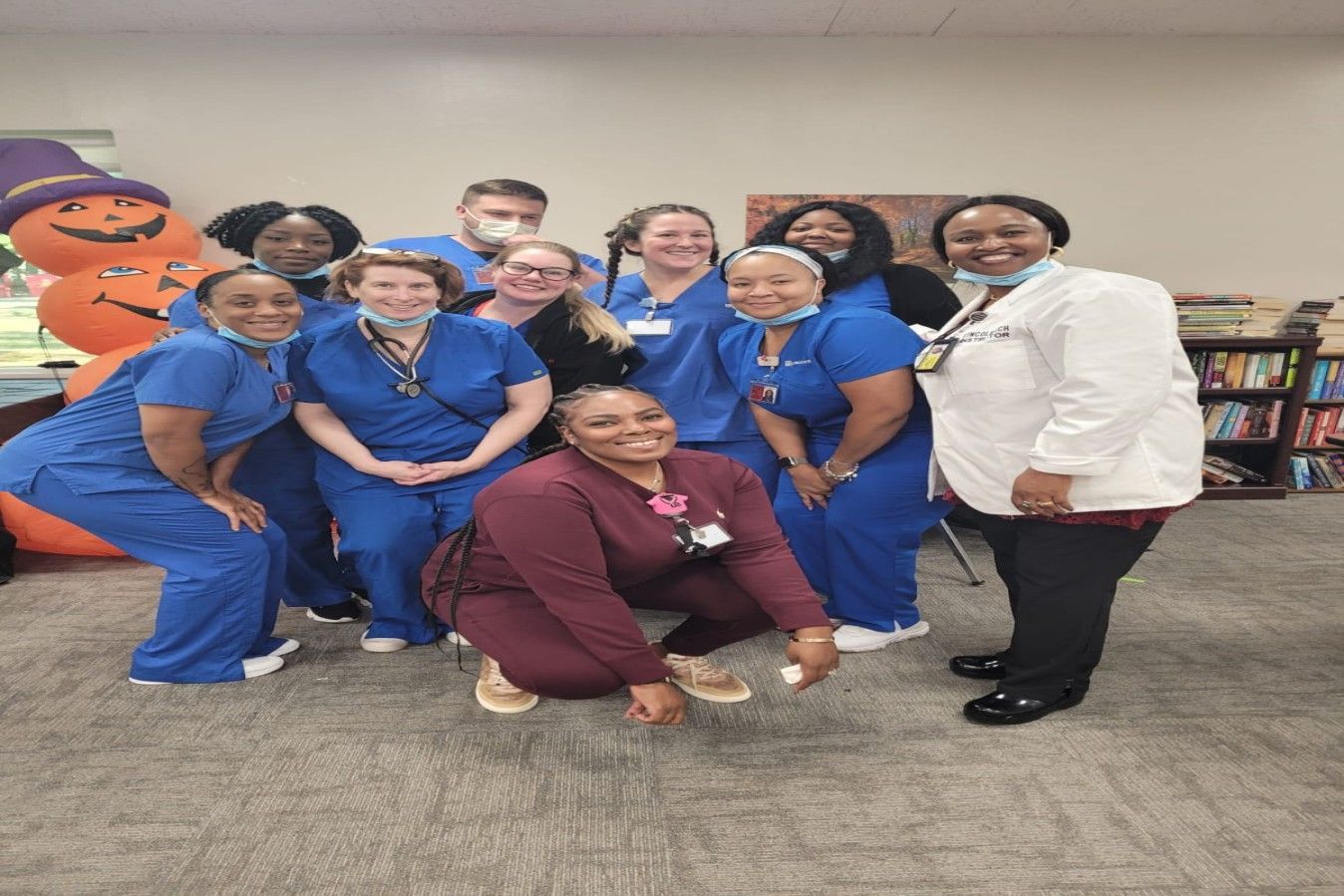 Group of nurses in blue scrubs and a white coat pose for a photo in a library with Halloween decorations.
