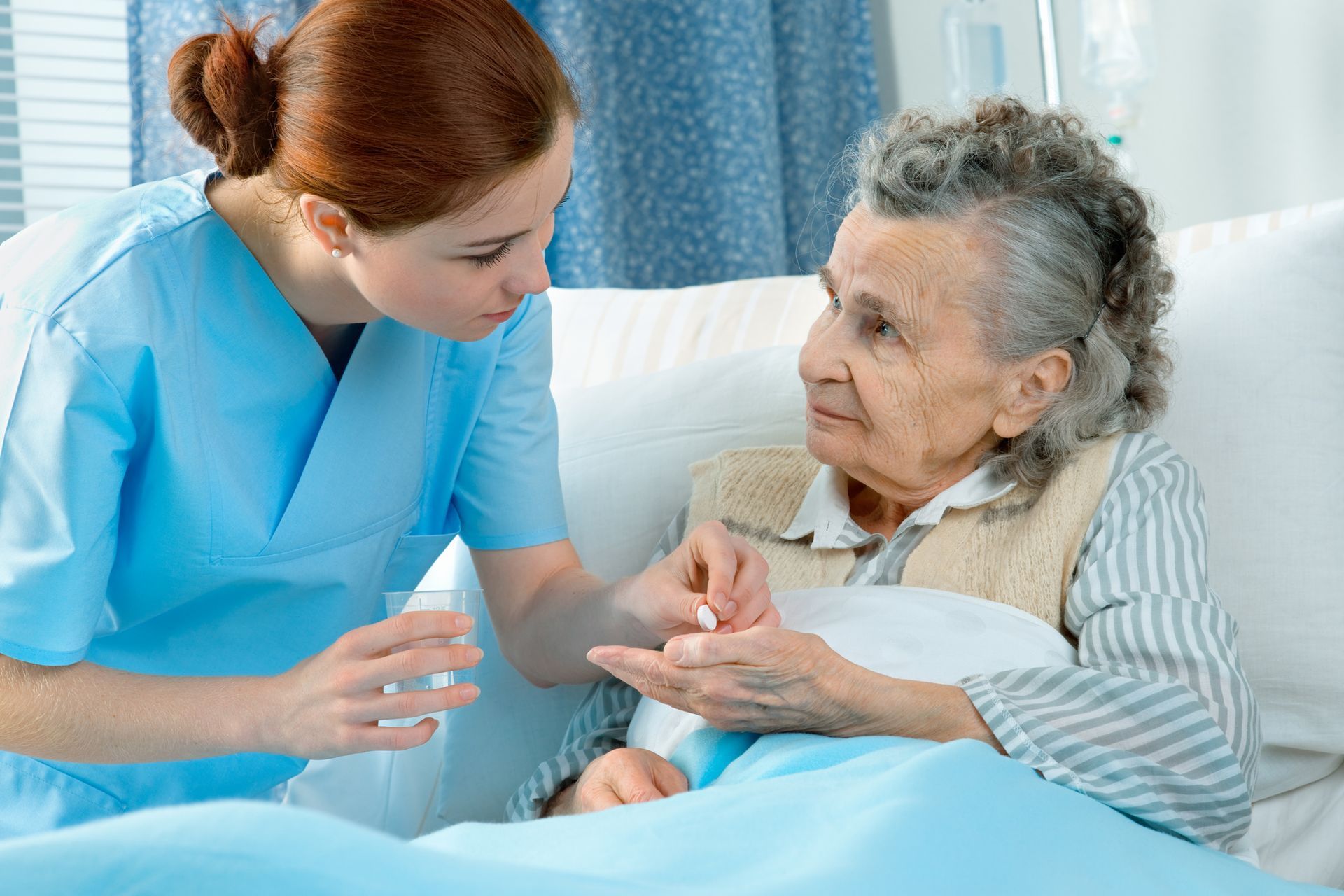 Nurse in blue scrubs giving medication to elderly woman in bed.