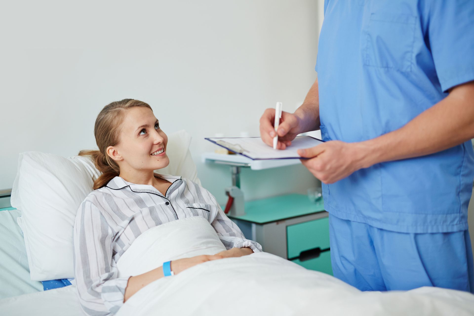 Woman in hospital bed smiles as healthcare worker in blue scrubs takes notes.