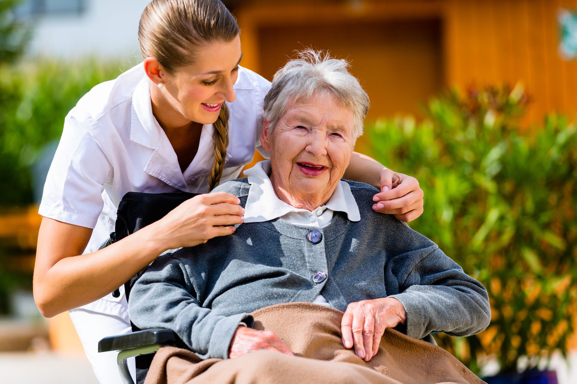 Woman in uniform assists elderly woman in a wheelchair outdoors; smiling faces, sunshine.