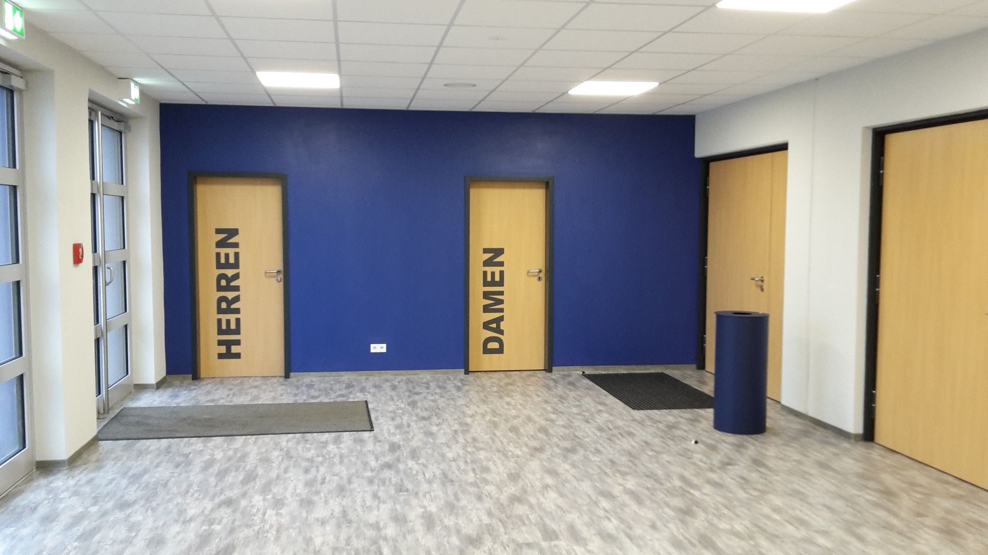 Ein Flur mit einer blauen Wand, an der sich zwei Holztüren mit der Aufschrift „HERREN“ (Männer) und „DAMEN“ (Frauen)