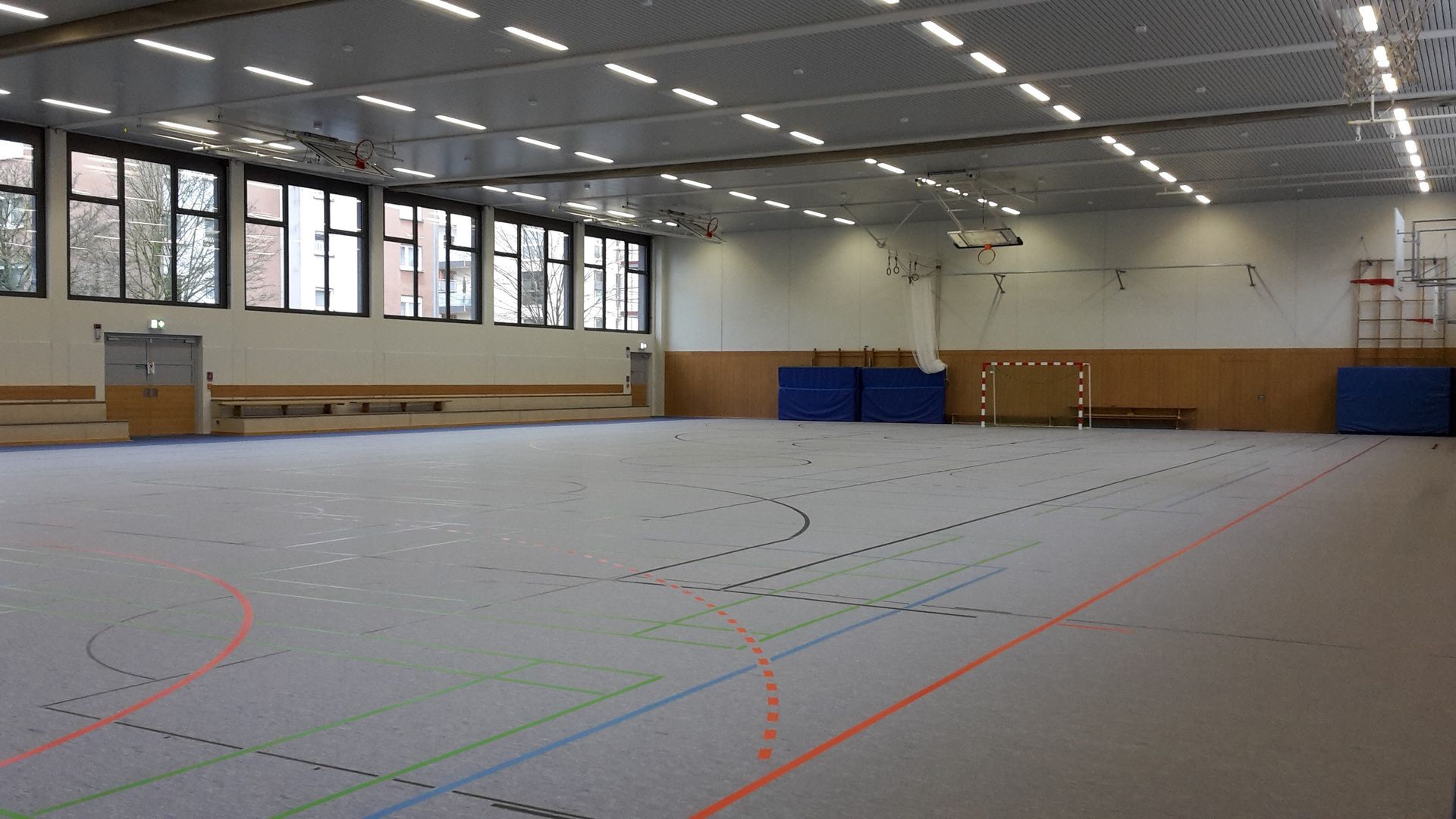 Eine geräumige Sporthalle mit hellem Boden, der mit verschiedenen farbenfrohen Sportfeldmarkierungen versehen ist, und 