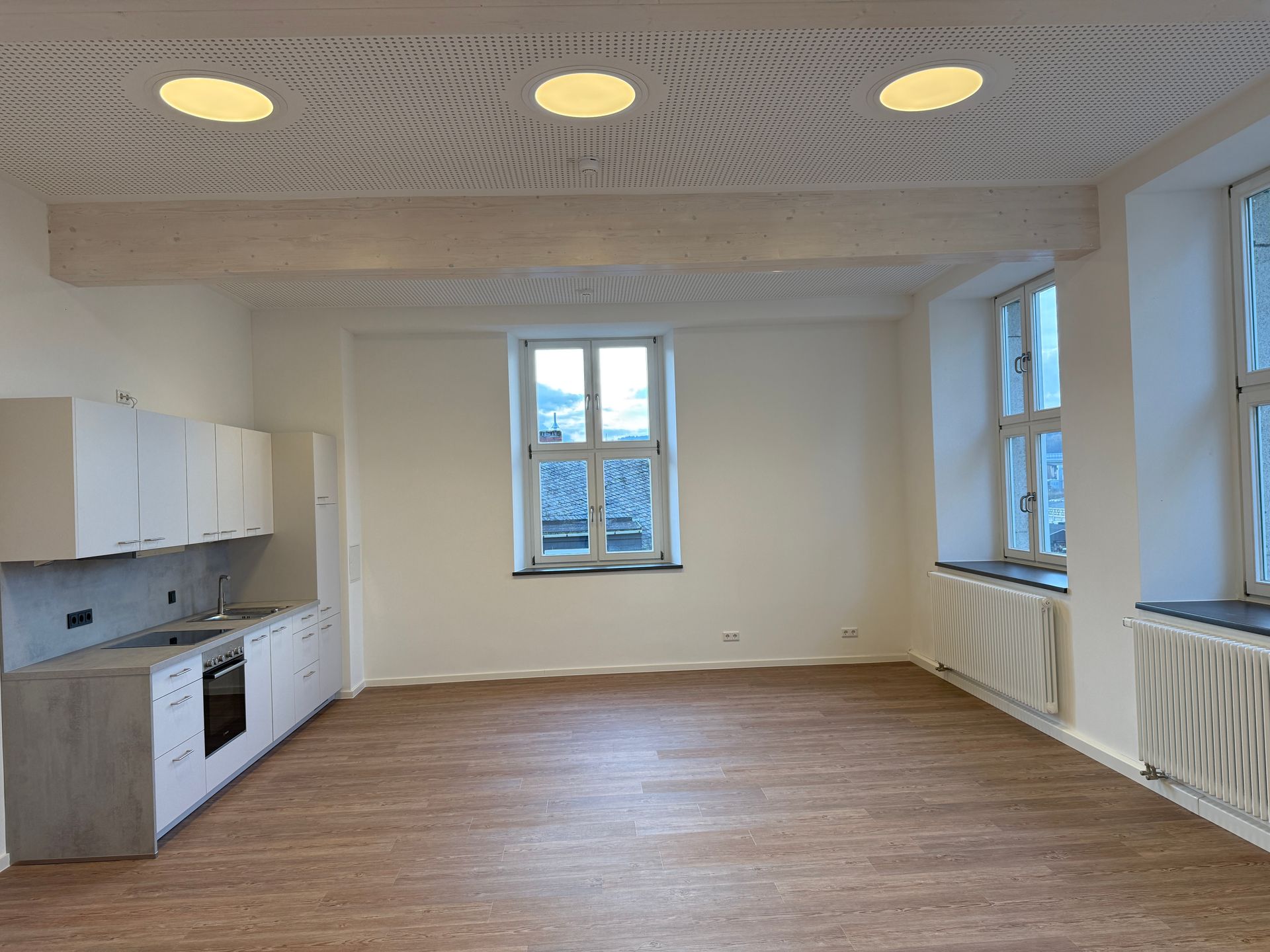 Eine leere, moderne Wohnung mit hellem Holzboden, weißen Küchenschränken und großen Fenstern mit Heizkörpern darunter.
