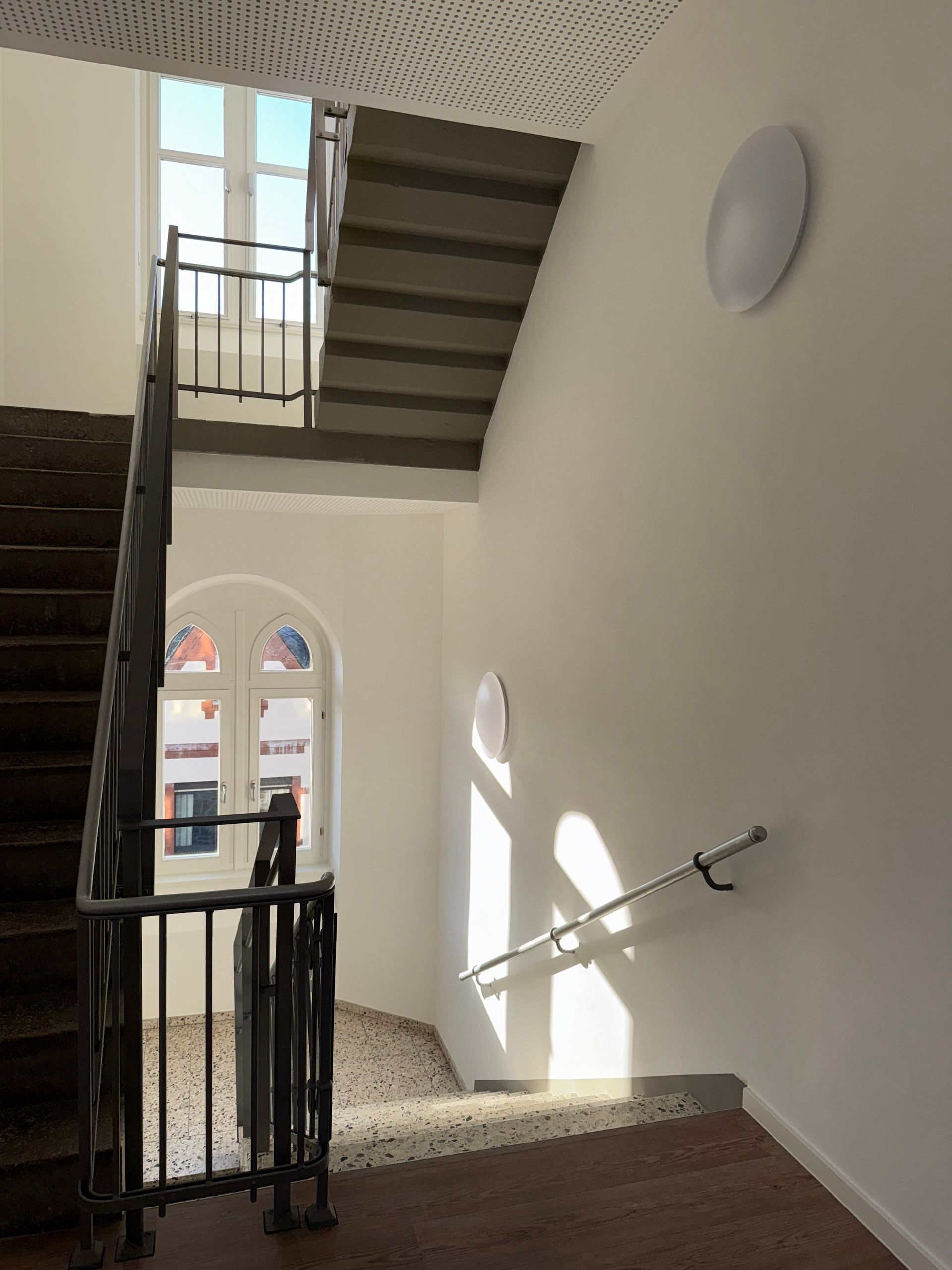 Eine mehrstufige Treppe mit dunklem Metallgeländer, weißen Wänden und einem großen Fenster mit Blick auf eine Straße.
