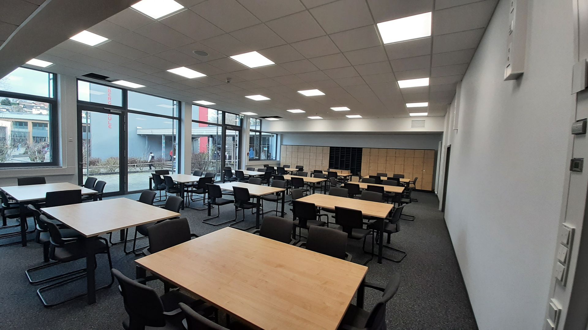 Ein leeres Klassenzimmer mit hellen Holztischen, schwarzen Stühlen, großen Fenstern und einem grauen Teppichboden.