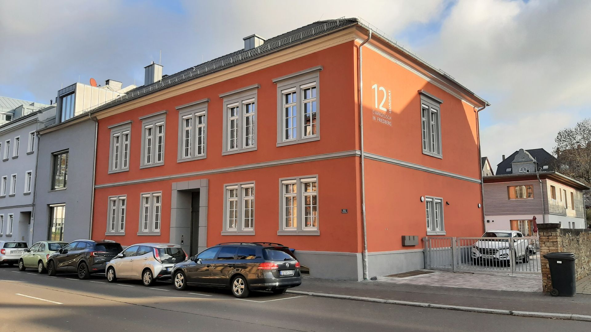Ein zweistöckiges, orangefarbenes Gebäude mit weiß gerahmten Fenstern an einer Straße, davor parken Autos.