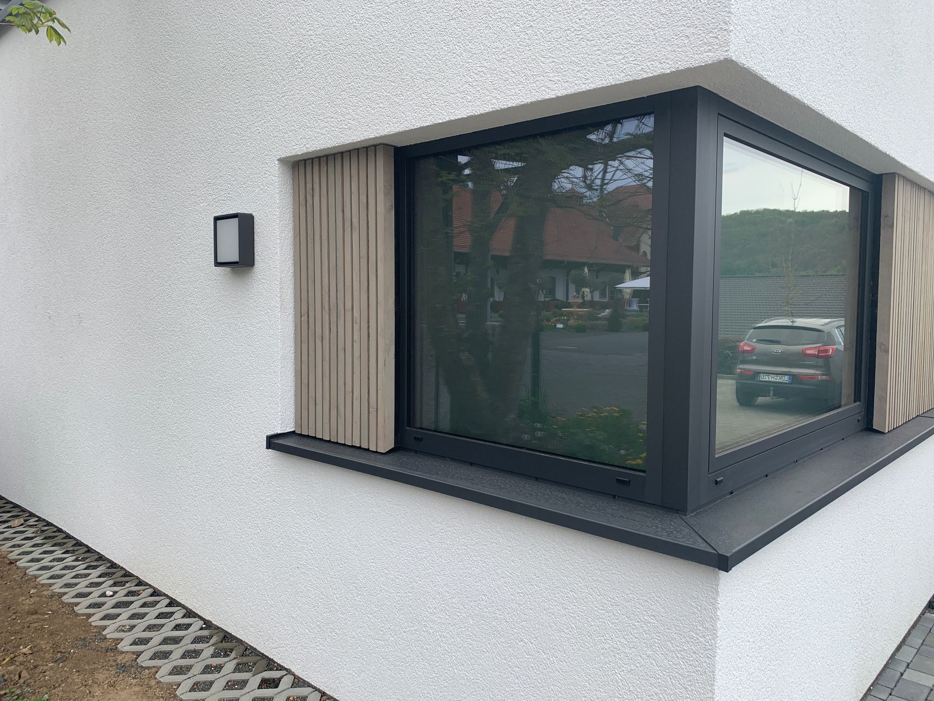 Eine weiße Stuckfassade mit einem schwarz gerahmten Eckfenster mit vertikalen beigen Lamellen und einer Außenwandleuchte.