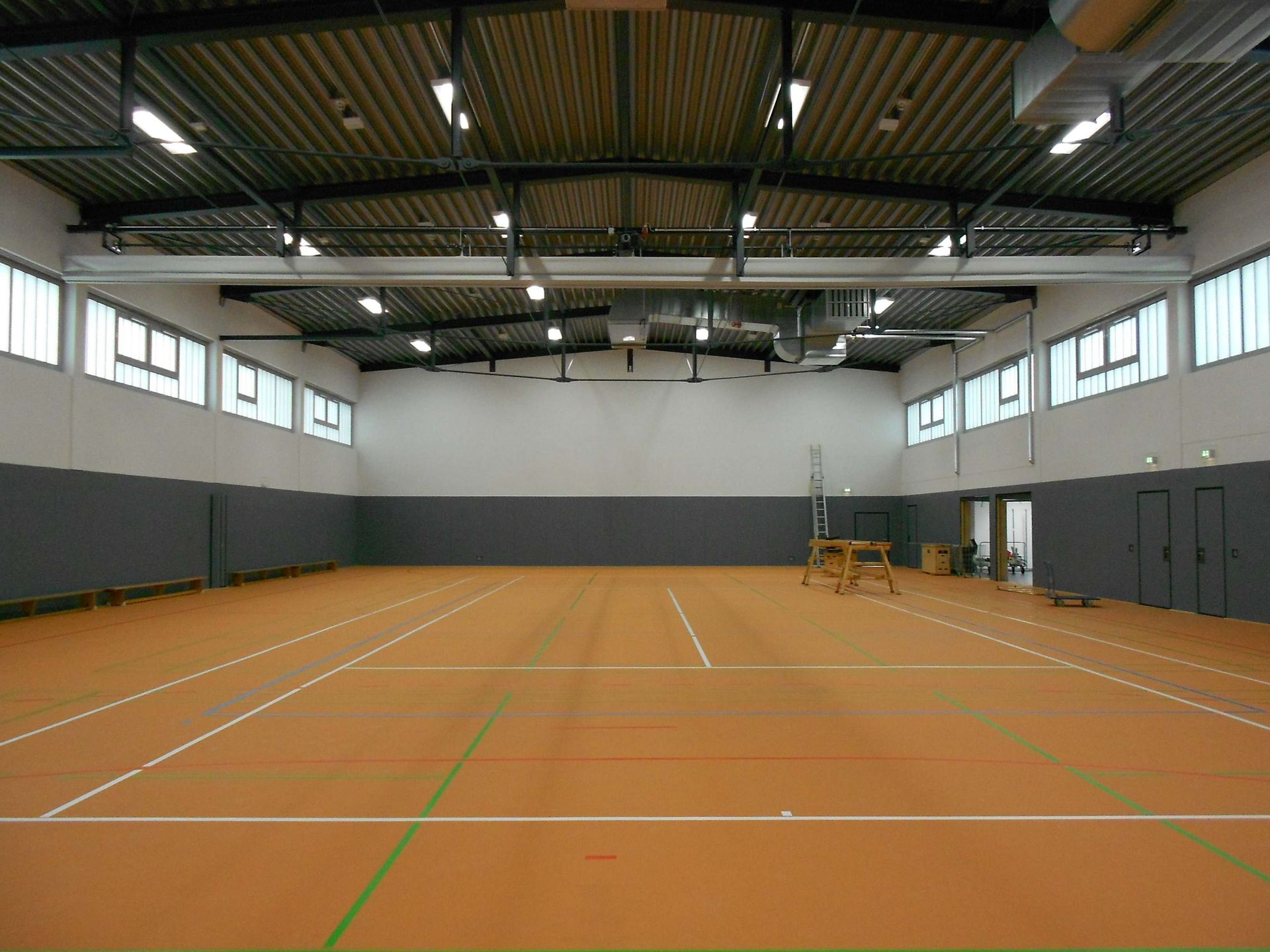 Eine Indoor-Sporthalle mit beigefarbenem Spielfeld, weißen Linien, grauem Wandsockel und hoher Decke mit Deckenbeleuchtung.